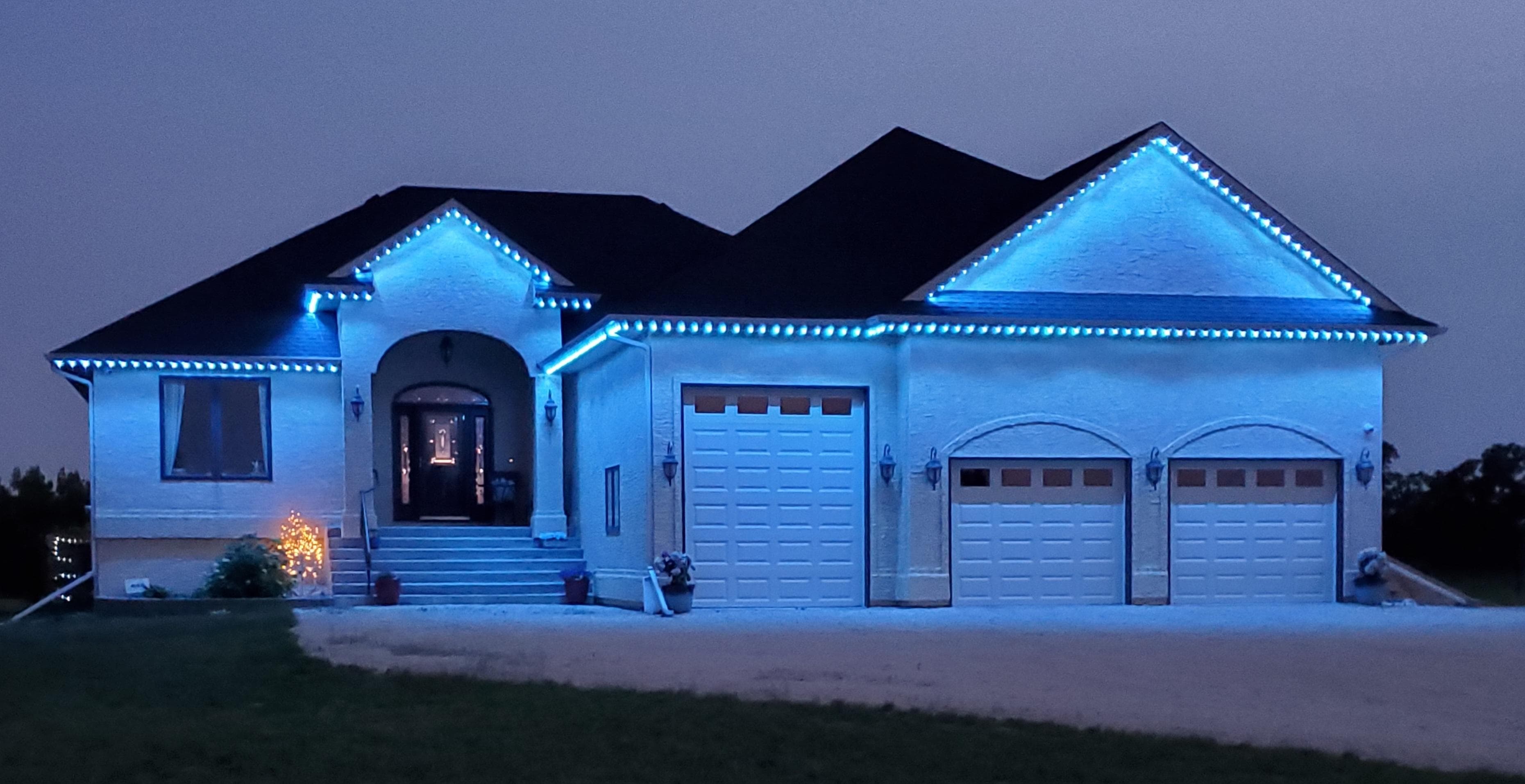 Two-story Burlington home with permanent blue LED outdoor lighting along every roofline, with the Halton Glow control app shown on a phone in the driveway